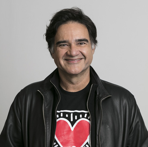 Sergio Valente com uma jaqueta preta e uma camisa preta com estampa de um coração e traços brancos sorrindo para a câmera em um fundo branco