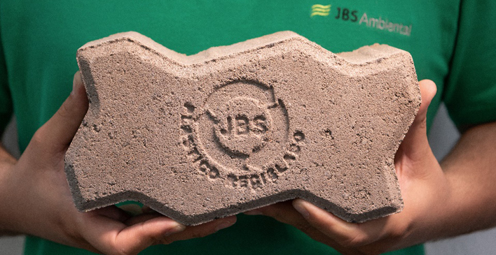 uma pessoa de camisa verde com o logo da JBS Ambiental segurando um tijolo com o logo da JBS e os dizeres plástico reciclado