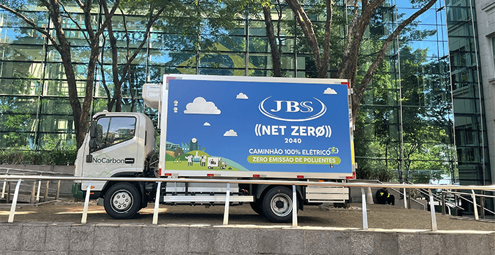 caminhão parado em frente a um prédio e duas arvores com logo no carbon na porta e em sua lateral escrito JBS Net Zero 2040 Camnihão 100% elétrico Zero emissão de Poluentes